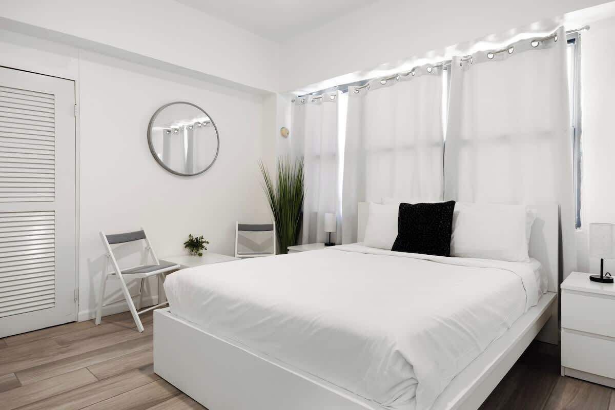 A modern bedroom with a large bed, a bedside table, a chair, a mirror, and a window with curtains.
