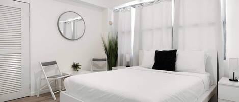 A modern bedroom with a large bed, a bedside table, a chair, a mirror, and a window with curtains.