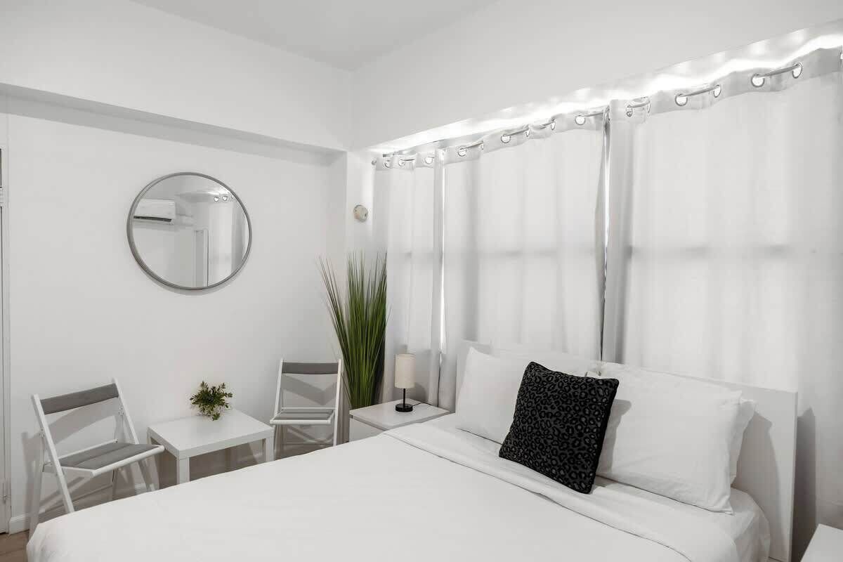 Et hotelværelse med en seng, et rundt spejl, et lille bord med en lampe, en stol og en potteplante.