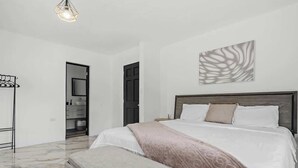 Un dormitorio con cama, una mesita de noche, una obra de arte colgada en la pared y una puerta que da al baño.