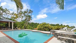 Una piscina con un juguete inflable verde, rodeada de palmeras y sillones de descanso.