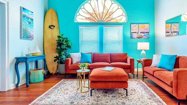 Een kamer met een blauwgroene wand, een groot raam met luiken, een surfplank, een rode bank, een klein rond tafeltje en een potplant.