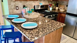 Een keuken met granieten aanrechtbladen, houten kastjes, een roestvrijstalen fornuis en blauwe barkrukken.