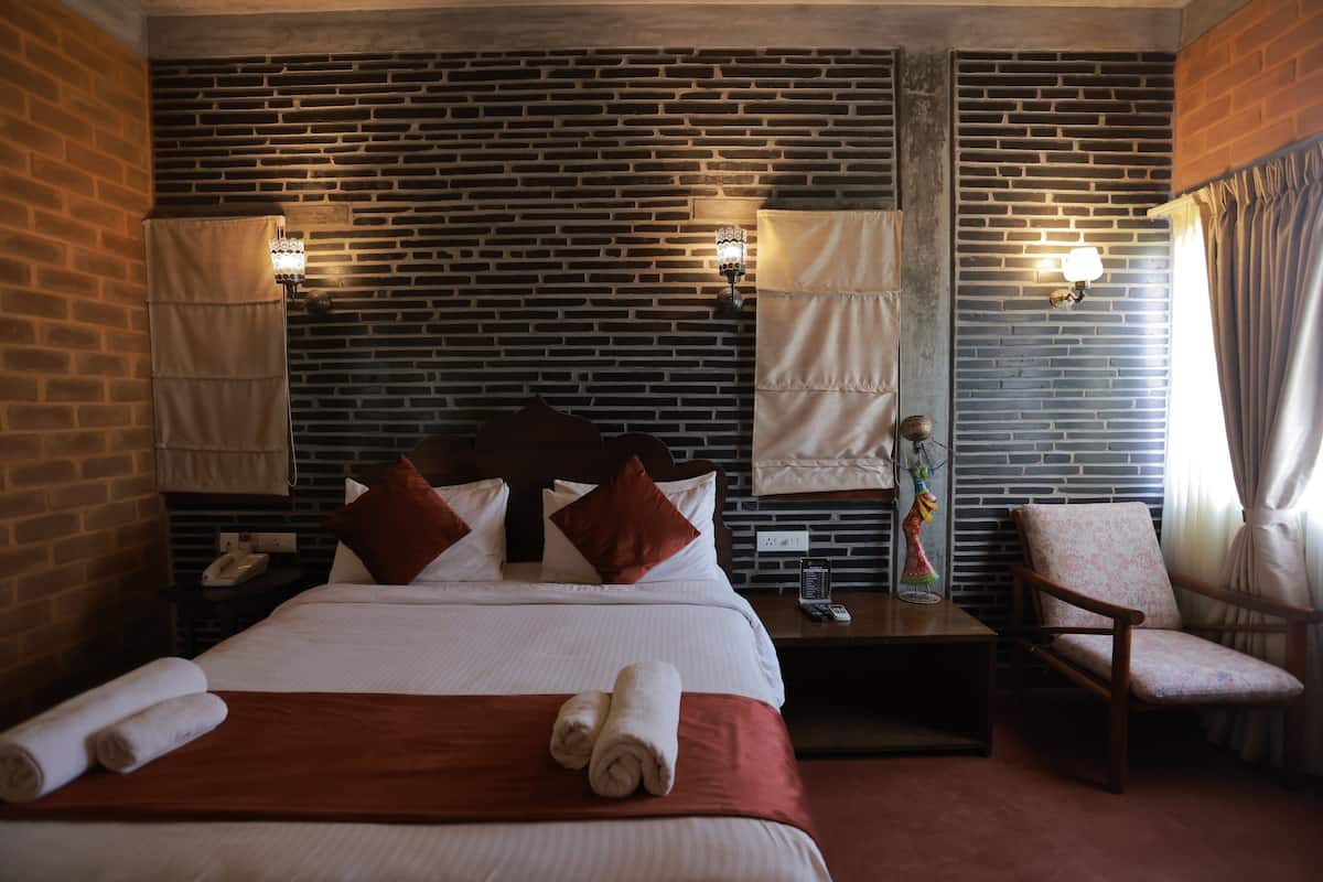 Une chambre d’hôtel avec un mur décoré d’un motif en briques, un grand lit avec une literie blanche, deux lampes de chevet, une table de chevet avec un téléphone, une chaise et une fenêtre avec des rideaux.