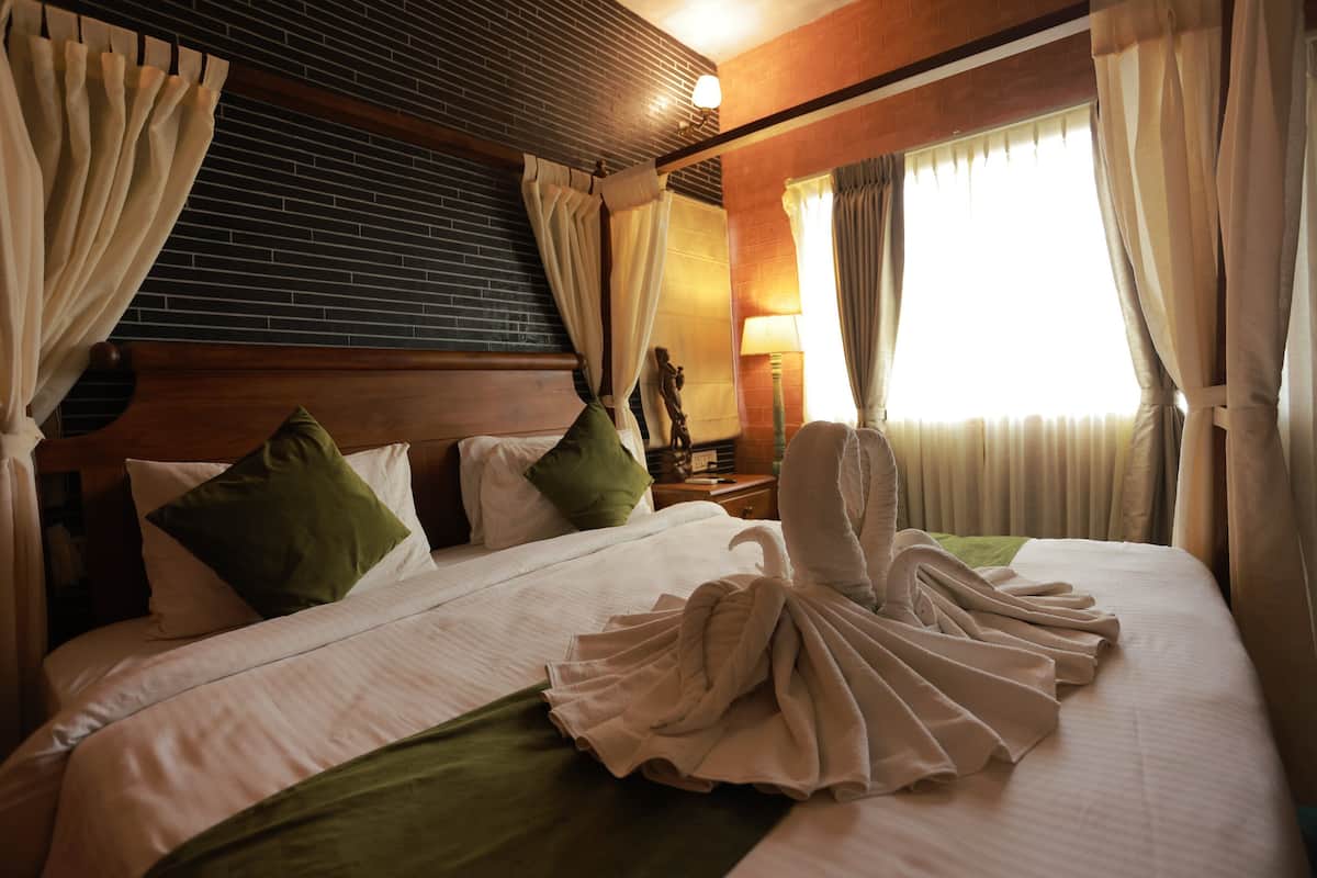 Une chambre d’hôtel avec un lit bien fait, une tête de lit en bois, une table de chevet avec une lampe et des rideaux aux fenêtres.
