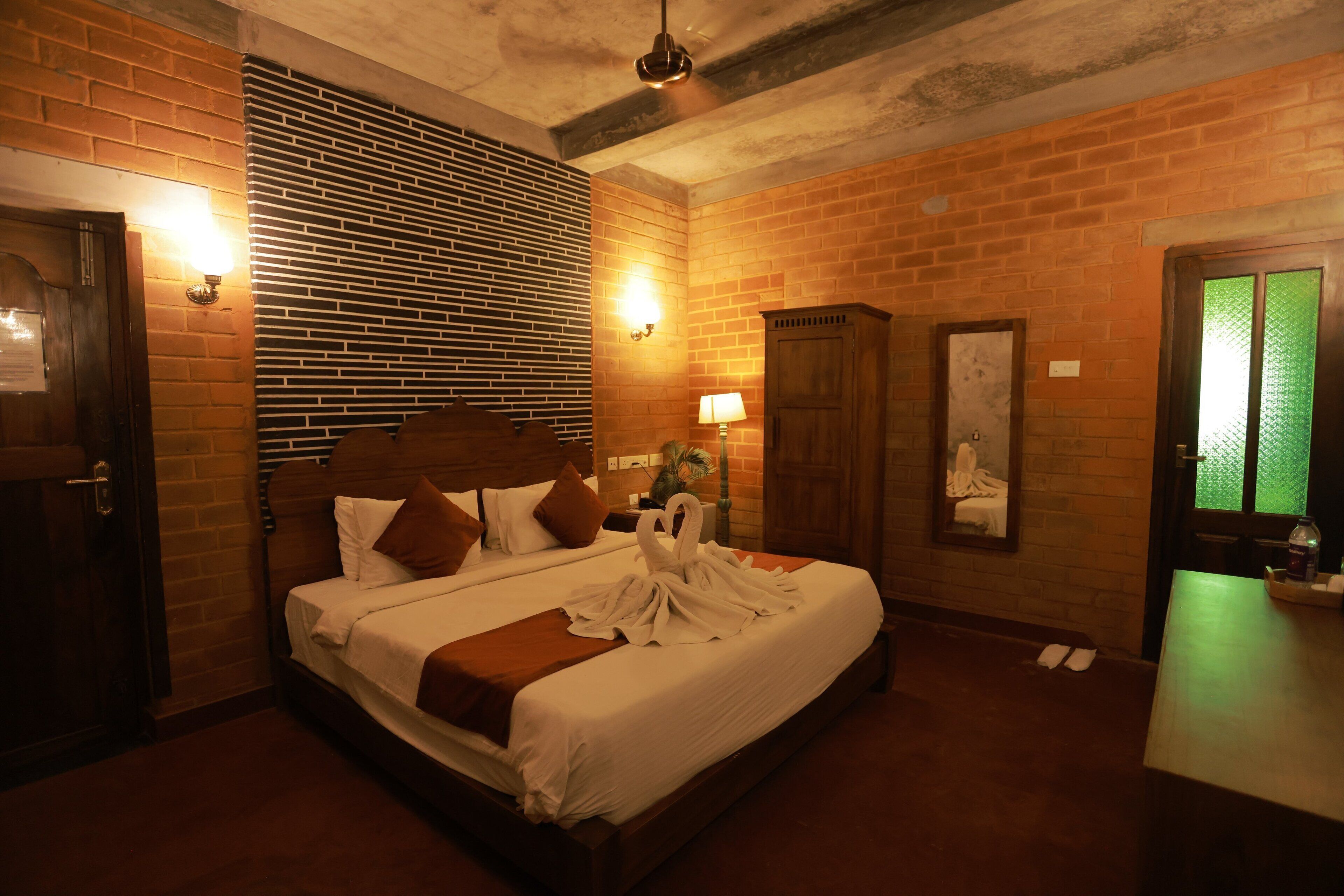 Una habitación de hotel con pared de ladrillo, cama de madera, mesita de noche, lámpara y espejo.