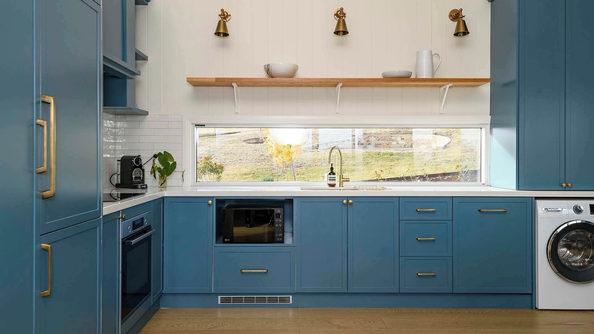 A modern kitchen with blue cabinets, a built-in oven, a washing machine, and a window with a view of greenery.