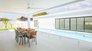 A modern outdoor dining area with a pool, white walls, and a clear view of the sky.