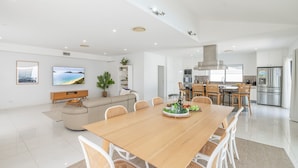 A modern, well-lit living room with a large wooden dining table, white wicker chairs, a beige sofa, and a flat-screen TV. - 57 Tomaree Road House - Pool, Air-con, Wi-fi, Boat Parking (Shoal Bay)