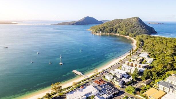 Vista aerea di una zona costiera con spiaggia, barche e area residenziale con edifici e vegetazione.