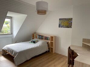 Une chambre avec un plafond en pente, un lit recouvert d’une courtepointe blanche, une bibliothèque en bois, un bureau avec une chaise et une fenêtre donnant sur un paysage verdoyant.