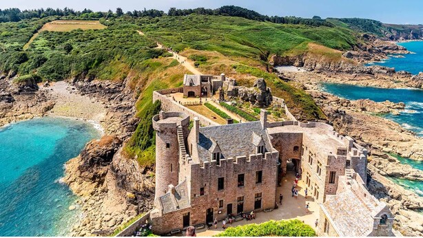 Eine historische Steinburg mit Innenhof in der Nähe einer felsigen Küste und üppigen grünen Hügel.