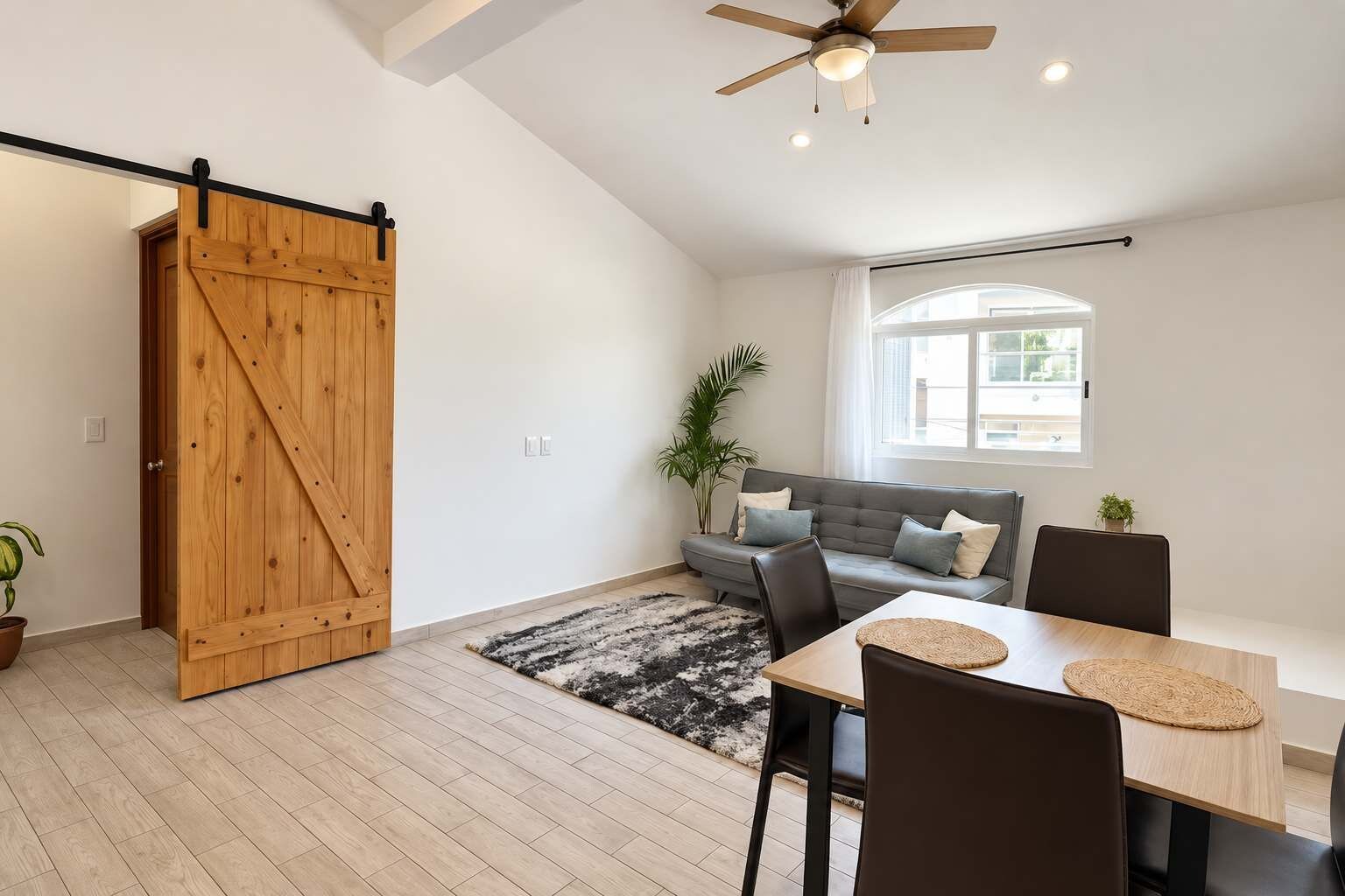Een moderne woonkamer met een schuifdeur in boerderijstijl, een grijze bank, een eettafel met stoelen en een plafondventilator.