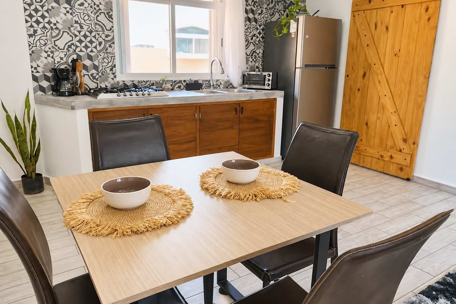 Un comedor con una mesa dispuesta para dos, una cocina con refrigerador y tostadora, y una planta en maceta.