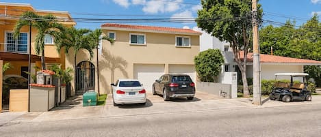 Una calle residencial con dos coches aparcados frente a una casa color beige con tejado de tejas rojas.