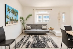 Ein modernes Wohnzimmer mit einem grauen Sofa, einem schwarz-weißen Teppich, einem Esstisch mit Stühlen, einer Pflanze und einem Palmen-Gemälde.