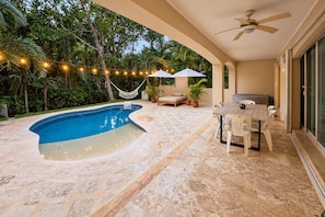 A swimming pool with a curved edge, surrounded by a patio area with a table and chairs, and a ceiling fan above. - 7BR Villa • Private Pool + Hot Tub • Beach 5 Min (Puerto Aventuras)