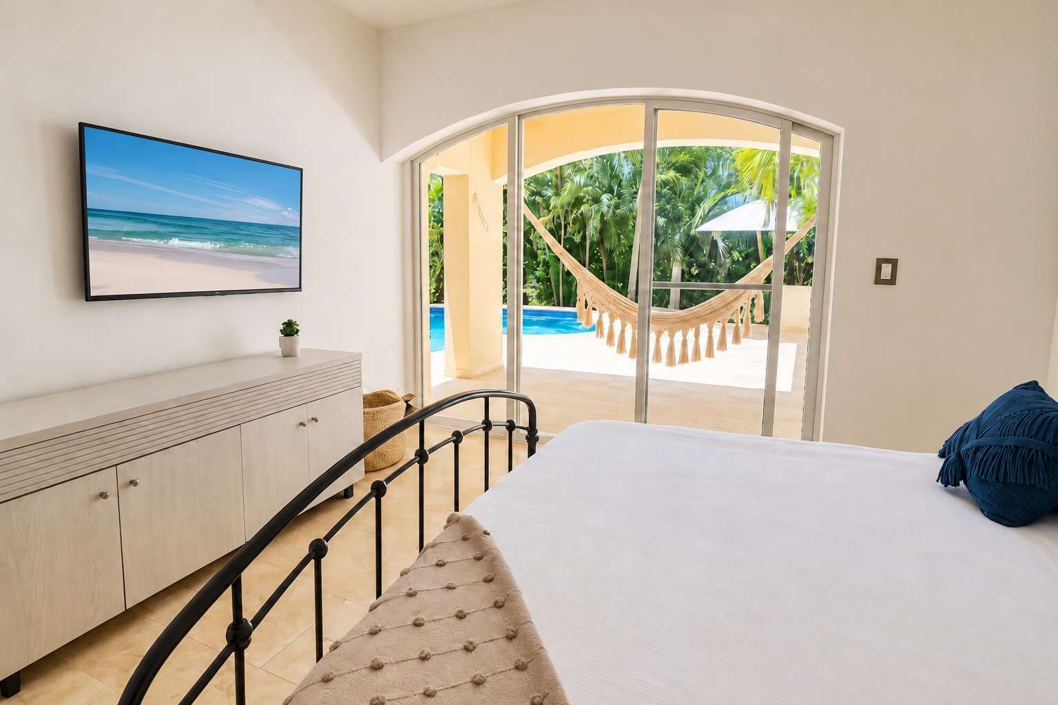 A bedroom with a bed, a TV on the wall, a view of a pool and palm trees through a glass door.