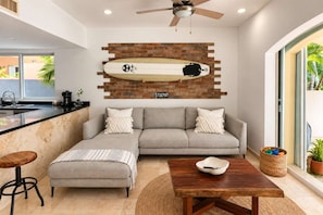 A modern living room with a sofa, ottoman, coffee table, and a surfboard mounted on a brick wall. - 7BR Villa • Private Pool + Hot Tub • Beach 5 Min (Puerto Aventuras)