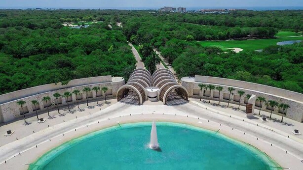 Um resort com piscina central, palmeiras e um campo de golfe ao fundo.