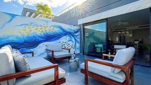Área externa com assentos, mobiliário moderno, um mural de onda e peixes, e vista para o interior do edifício.
