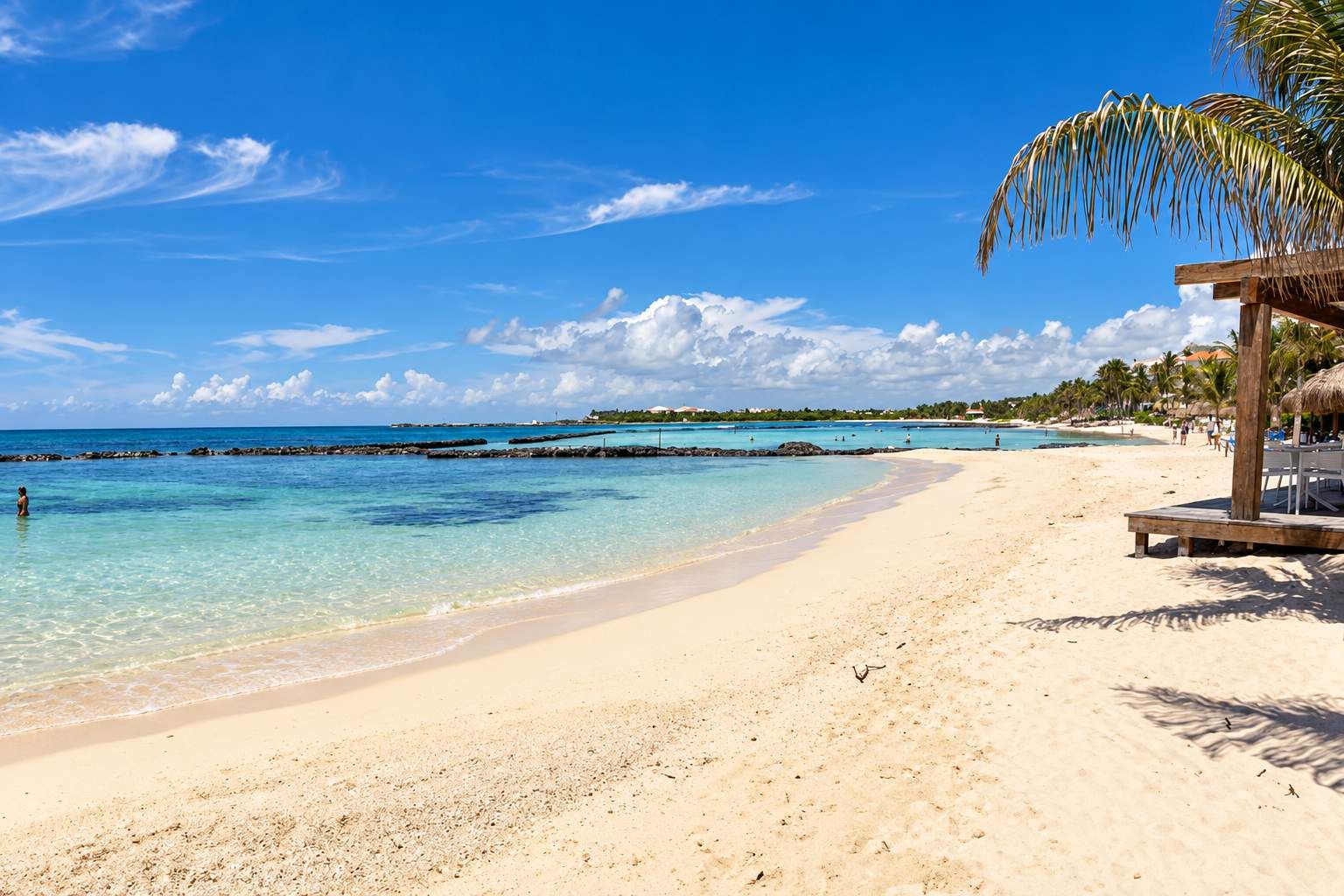 Une plage avec une eau turquoise et limpide, un rivage sablonneux et une structure en bois surmontée d’un toit de chaume.