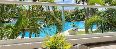 Un balcón con barandilla de vidrio que da a una piscina rodeada de palmeras y un cielo despejado.