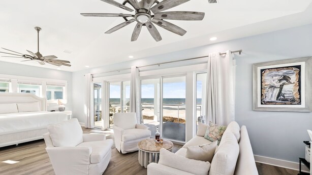 Ein geräumiges Wohnzimmer mit großem Fenster zum Strand, Deckenventilator und einer modernen weißen Sofagarnitur.