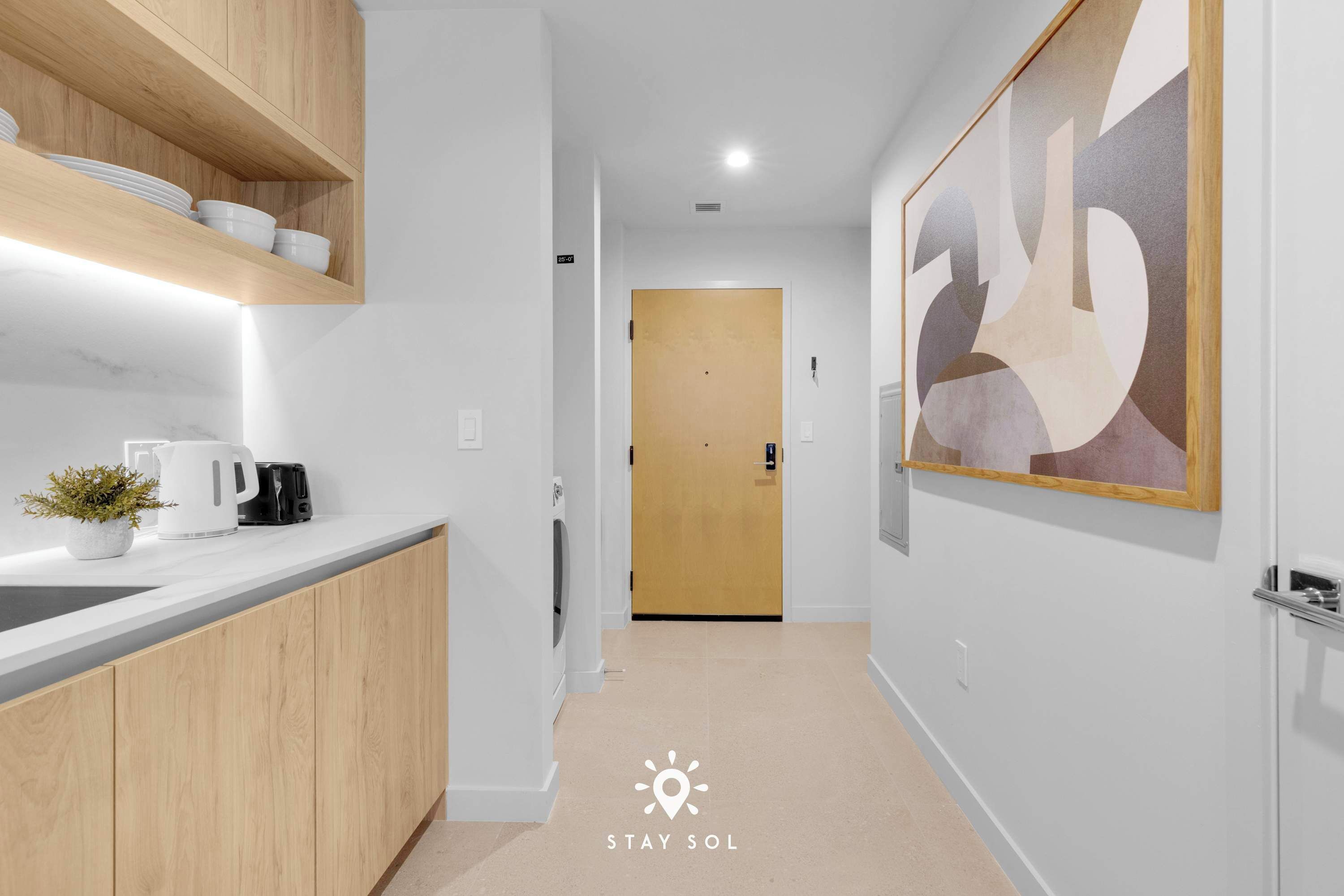 A modern kitchen with wooden cabinets, a countertop, and a coffee maker. There is a hallway with a door at the end and a framed artwork on the wall.