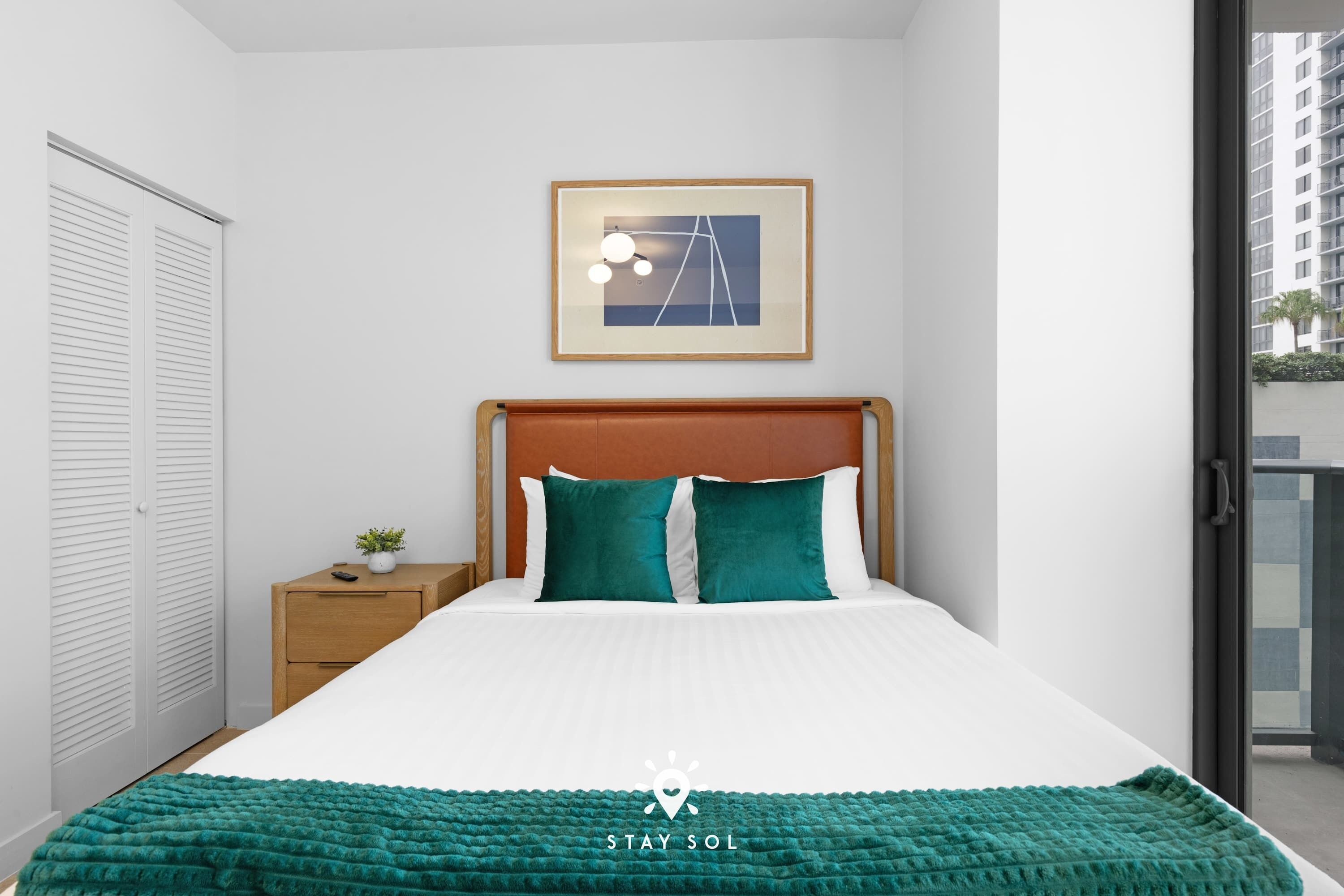 A neatly arranged bedroom with a bed, a nightstand, a framed picture, and a view of the outside balcony.