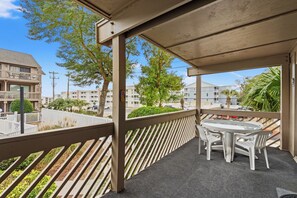 A covered outdoor area with a table and chairs, a wooden railing, and a view of surrounding buildings and greenery. - Steps to the Beach, Close to It All — Shipwatch II 101K (Myrtle Beach)