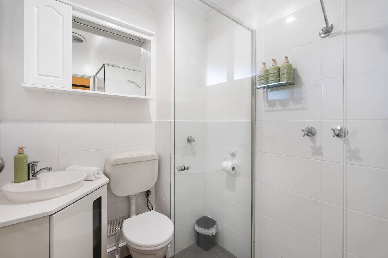 Un bagno moderno con lavandino, water e doccia bianchi. Sopra il lavandino c'è uno specchio e uno scaffale con articoli da toilette.