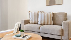 Un salon avec un canapé beige, des coussins à rayures, une petite plante et une télécommande posée sur une petite table ronde en bois.