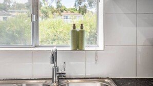 Une cuisine avec une fenêtre, un évier en acier inoxydable et deux bouteilles vertes étiquetées « Produits pour la vaisselle » posées sur le rebord de la fenêtre.
