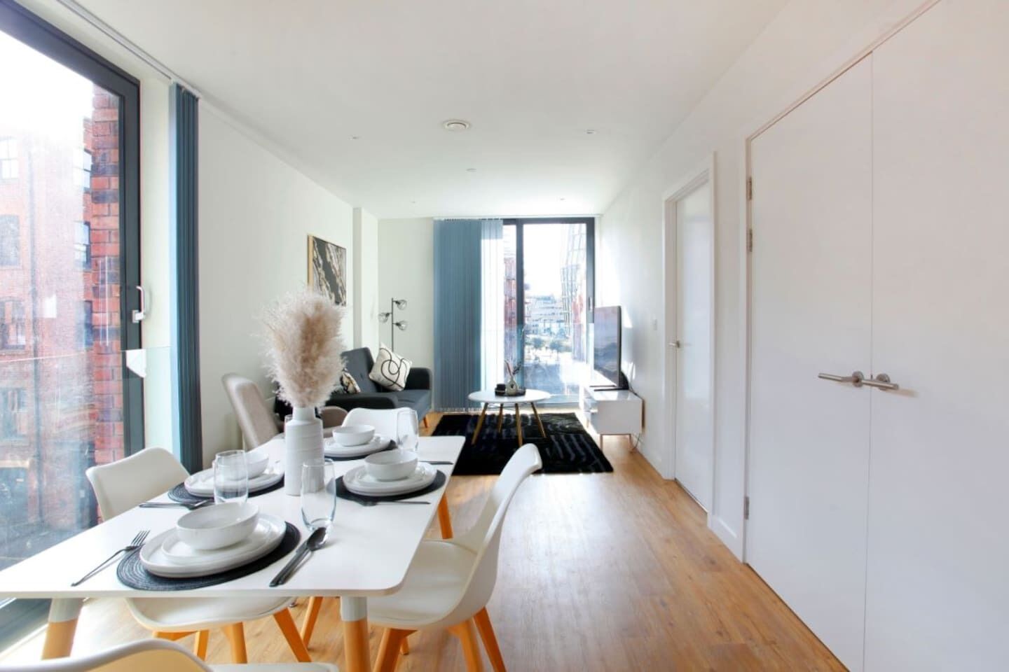 A modern dining area with a long table set for four, large windows, and a view of the city.