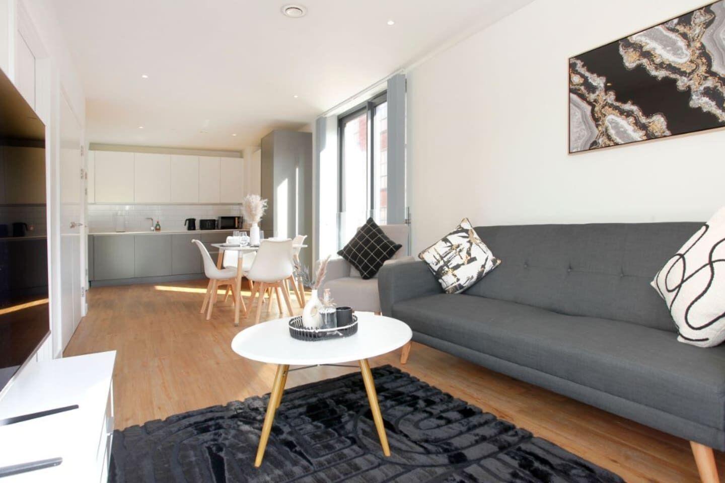 A modern living room with a grey sofa, a white coffee table, and a black rug. There is a dining area with white chairs and a table, and a kitchen in the background.