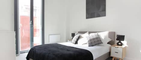 A modern bedroom with a large bed, a nightstand, a lamp, a ceiling light, and a view through a sliding glass door.