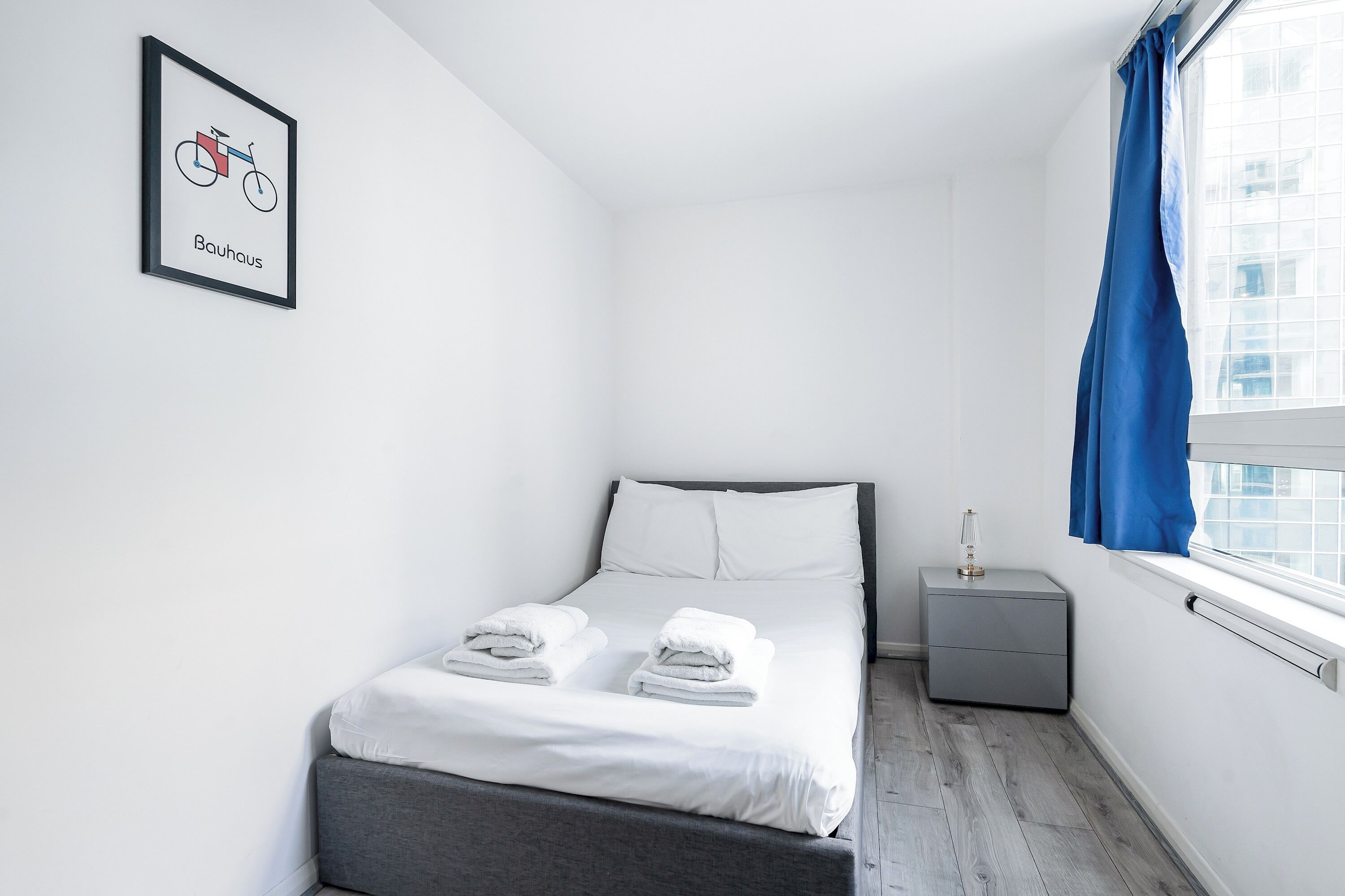 A minimalist hotel room with a bed, a nightstand, a framed picture, and a window with curtains.