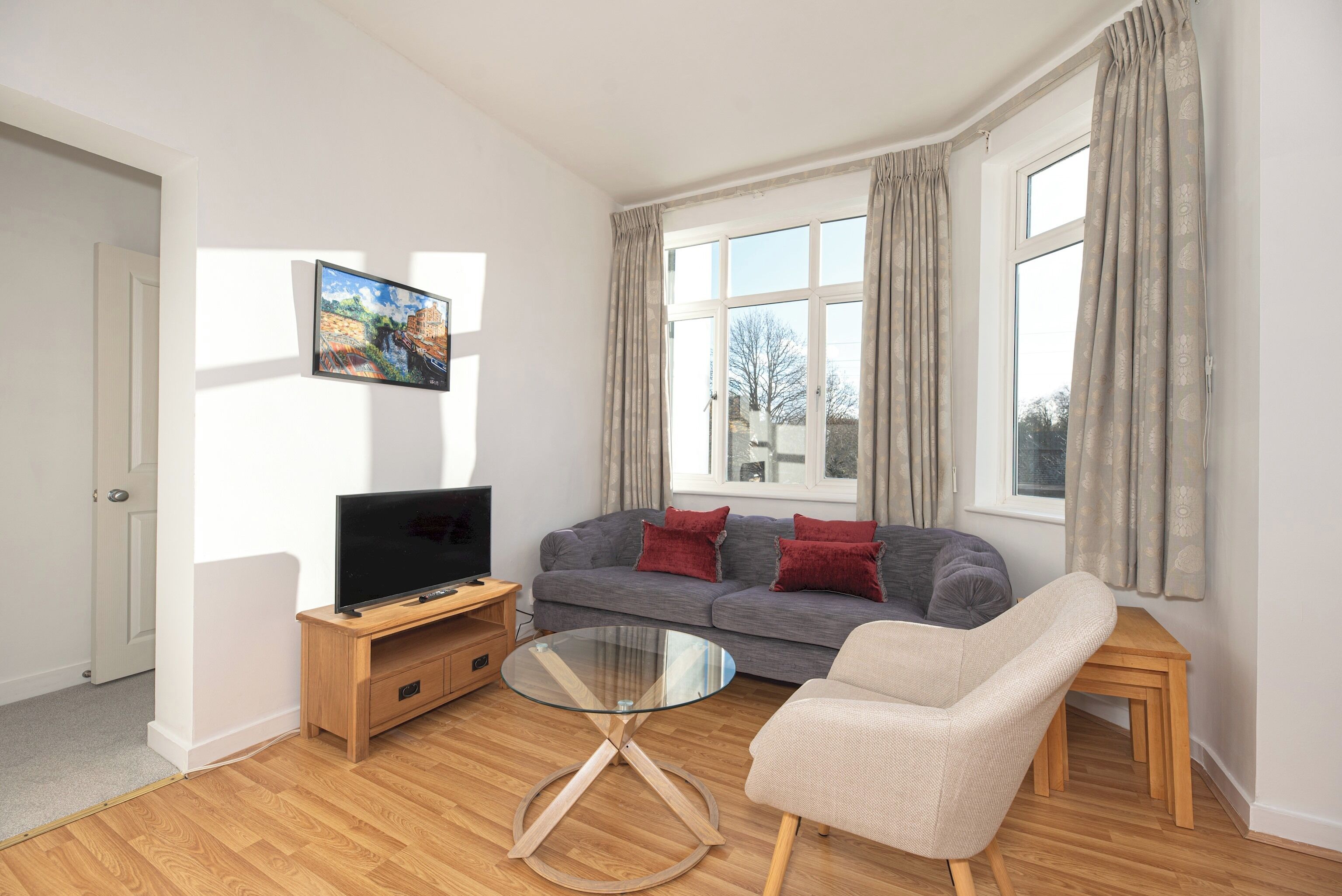 A modern living room with a grey sofa, a beige armchair, a glass coffee table, and a flat-screen TV on a wooden stand.