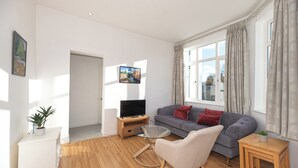 A modern living room with a grey sofa, a glass coffee table, a TV on a wooden stand, and a potted plant. - A beautiful 2-bed mansion-block flat in Putney - Pass the Keys (London)