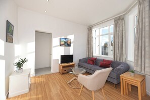 A modern living room with a grey sofa, a glass coffee table, a TV on a wooden stand, and a potted plant. - A beautiful 2-bed mansion-block flat in Putney - Pass the Keys (London)