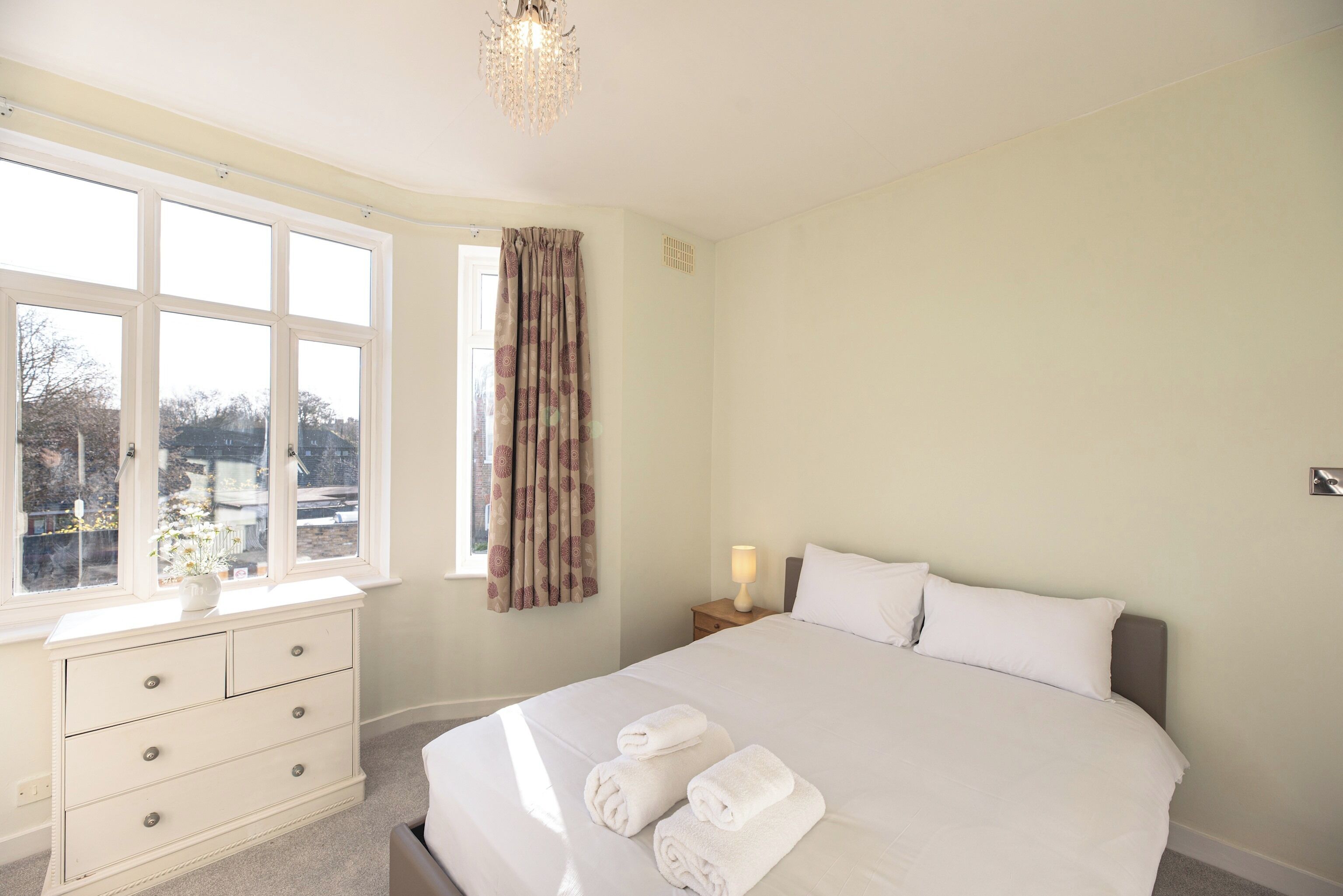 Une chambre à coucher avec un grand lit, une table de chevet, une commode et une fenêtre avec des rideaux.