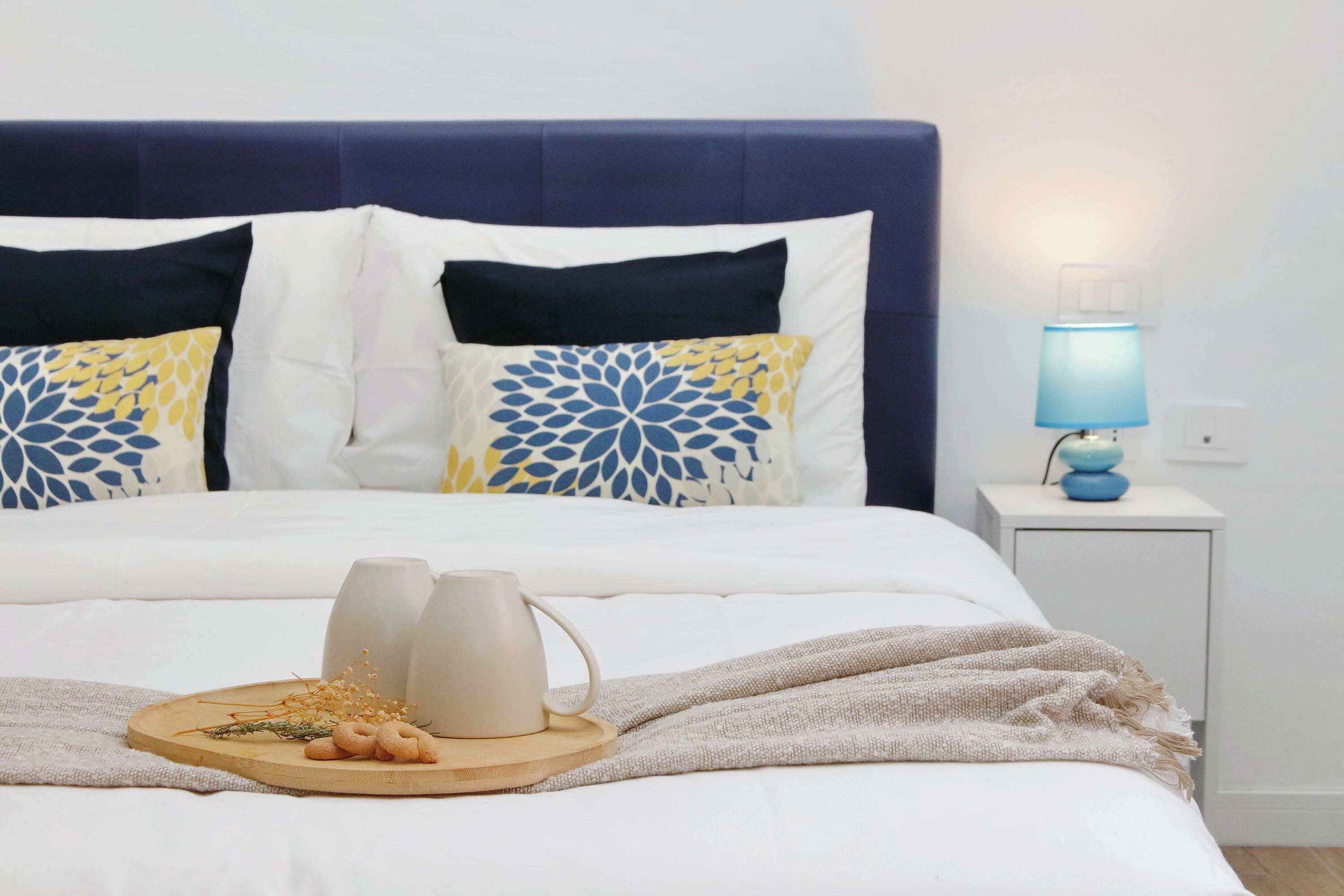 A neatly made bed with a blue headboard, white bedding, and decorative pillows. A bedside table with a lamp and a tray with candles and snacks is visible.
