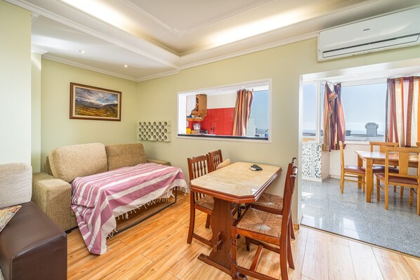 Ein Zimmer mit Esstisch, Stühlen, einer Couch, einem Bett mit gestreifter Decke, einem Bild an der Wand und Blick durch ein Fenster.