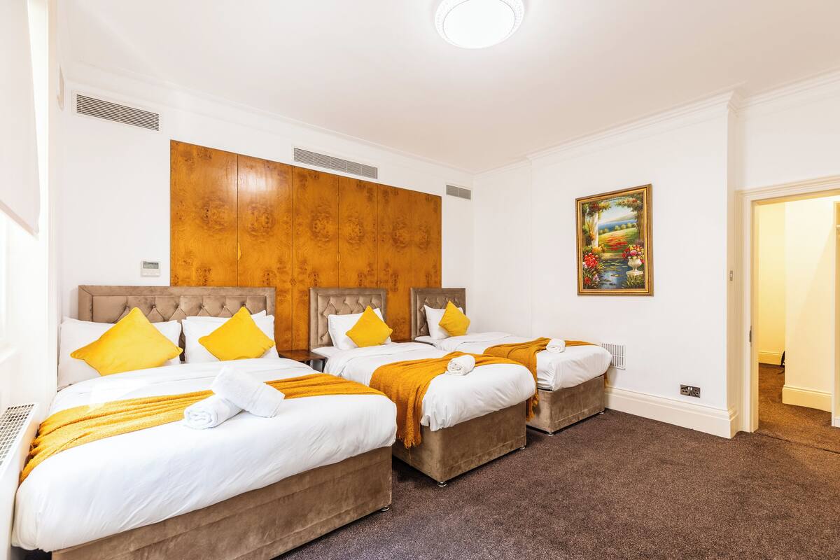 A hotel room with three beds, each with white bedding and yellow pillows. There is a wooden headboard, a painting on the wall, and a carpeted floor.