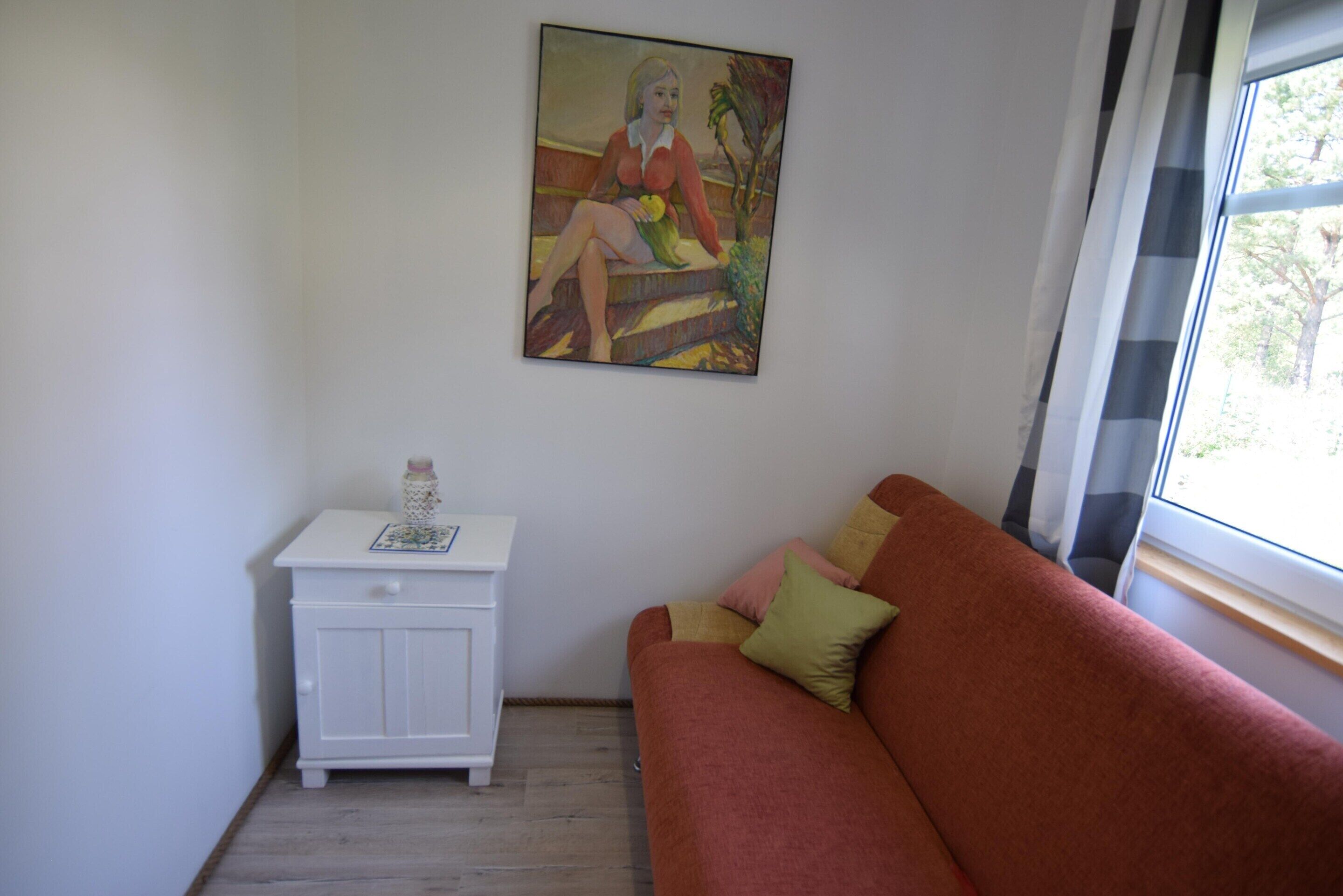 Ein Zimmer mit einem roten Sofa, einem weißen Nachttisch mit einer Flasche und einem Gemälde an der Wand.