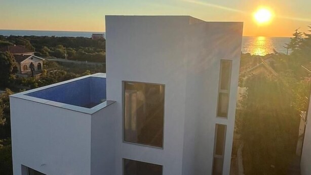 Ein modernes Gebäude mit flachem Dach und zahlreichen Fenstern, mit Blick auf eine Küstenlandschaft und Sonnenuntergang.