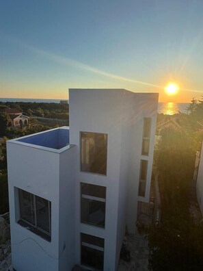 Un bâtiment moderne doté d’un toit plat et de nombreuses fenêtres, offrant une vue sur la côte et le coucher de soleil.
