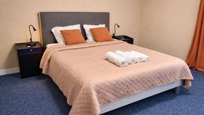 A neatly made bed with a quilted cover, two pillows, and rolled towels on a blue carpeted floor.