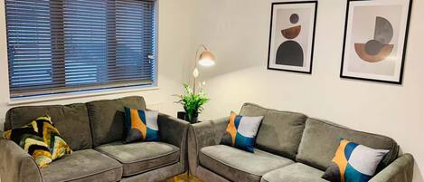 A living room with two grey sofas, a wooden floor, a window with blinds, a floor lamp, and two abstract wall art pieces.